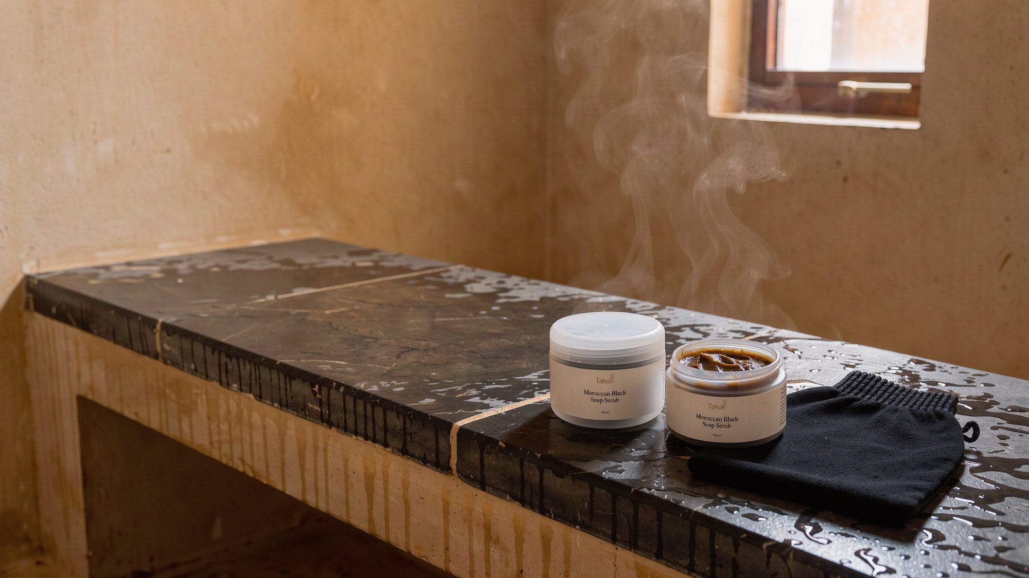 The black soap jar open on a stone bench in the hammam, a kessa glove beside it